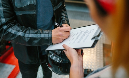 Motorcyclist in a leather jacket signing insurance claim paperwork while holding a helmet at an office counter.