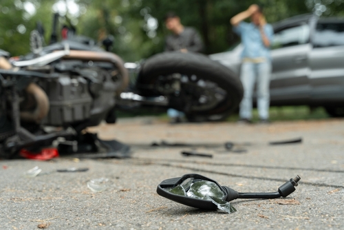 Close-up of shattered motorcycle mirror lying on the road after a serious collision, with a damaged bike and people in the background.
