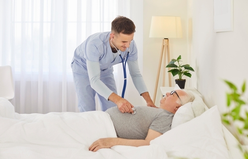 Nurse using a stethoscope to check an elderly male patient lying in bed.